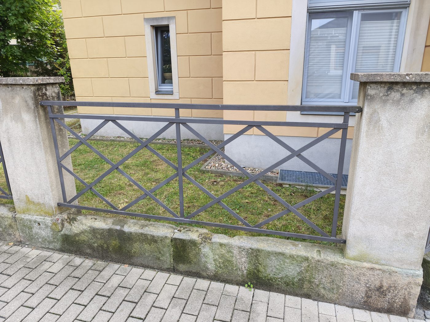 Metallzaun mit Rautenmuster zwischen Betonpfeilern vor einem Vorgarten